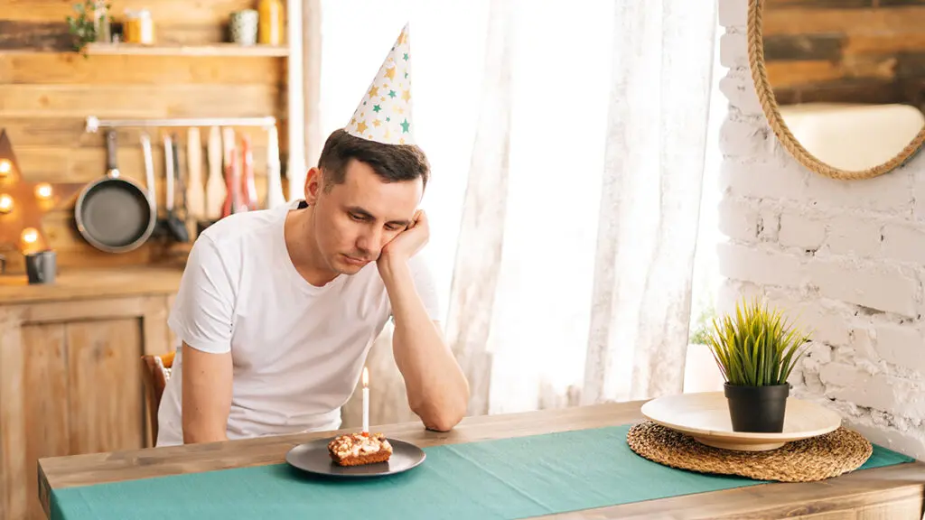 Upset young man sitting at the birthday cake and looking with sad eyes on it.