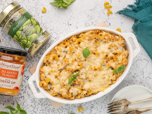 Artichoke mac n cheese in a casserole dish with two jars of dip next to it.