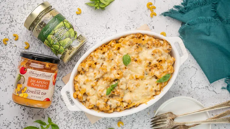 Artichoke mac n cheese in a casserole dish with two jars of dip next to it.