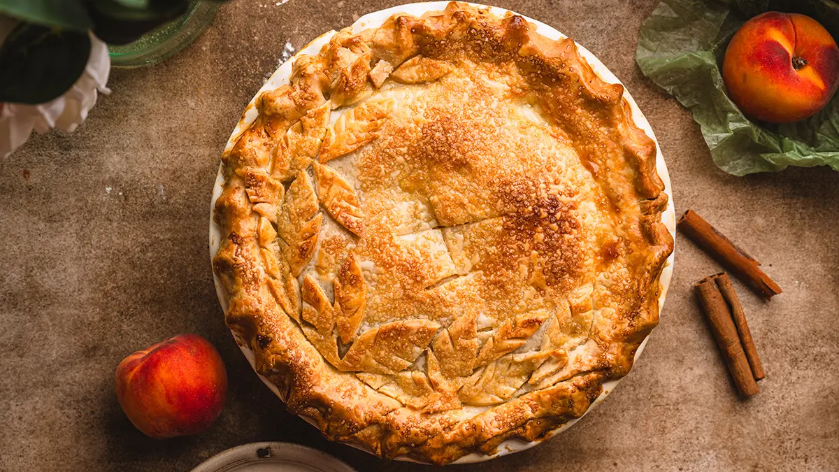 Peach pie on a table surrounded by a couple of peaches.