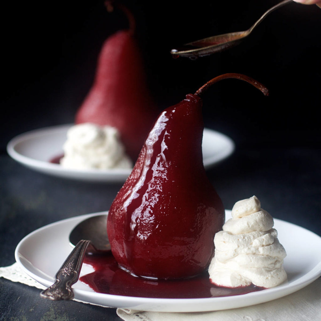 A photo of poached pears with two Spiced Red Wine Poached Pears on two plates with whipped cream.