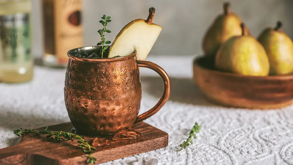 Moscow Mule with a slice of pear as garnish with pears and bottles in the background.