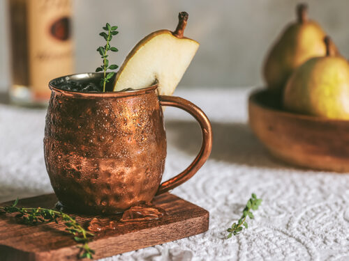 Moscow Mule with a slice of pear as garnish with pears and bottles in the background.