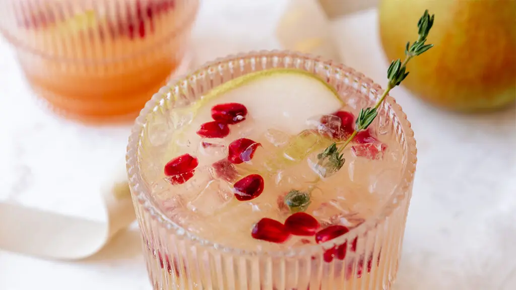 Pear kombucha mocktail in a glass with a slice of pear, pomegranate seeds, and thyme.