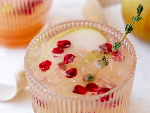 Pear kombucha mocktail in a glass with a slice of pear, pomegranate seeds, and thyme.