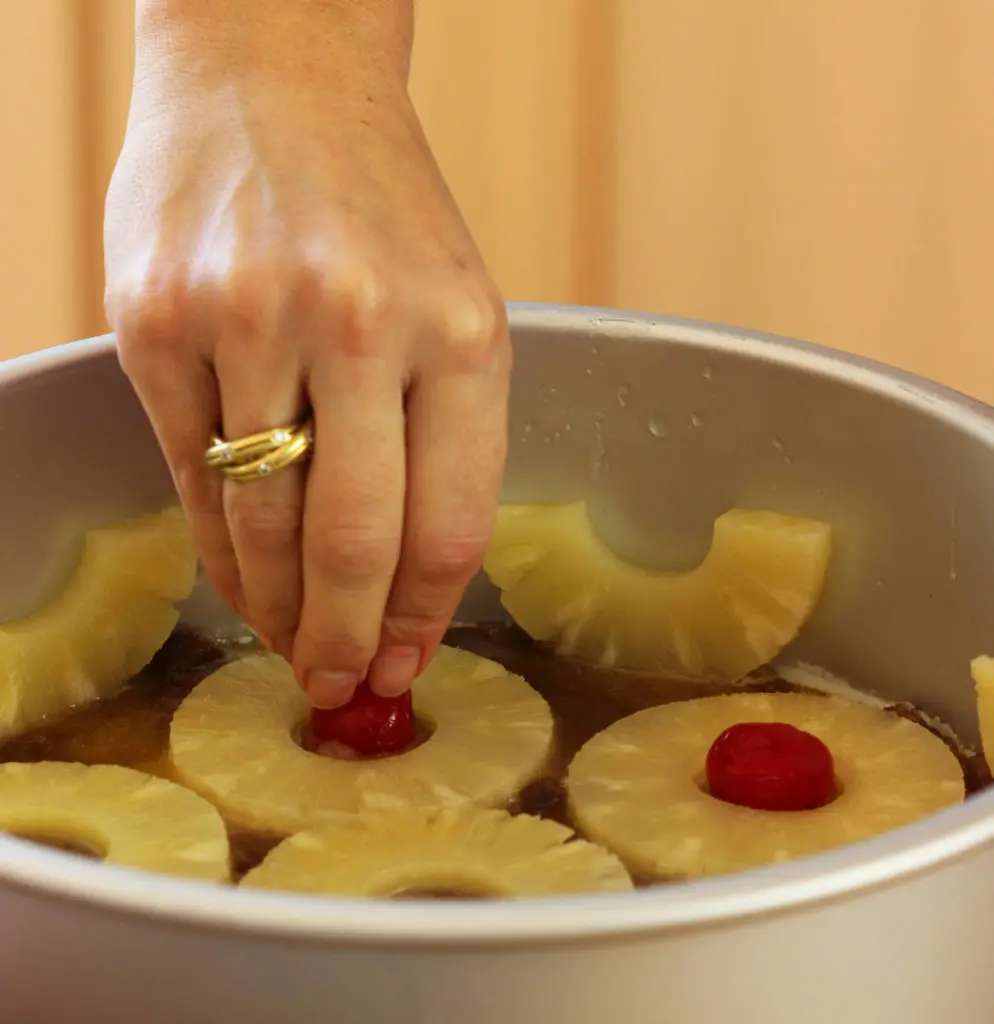 Pineapple upside down cake with a hand placing maraschino cherries in pineapple slices.