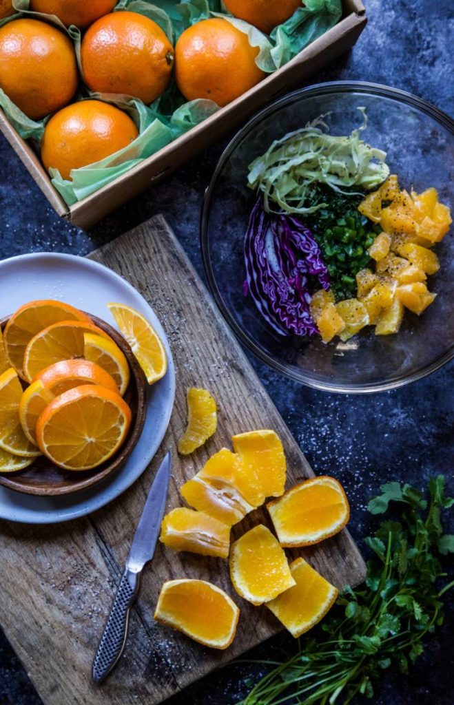 A photo of a salmon recipe with a box of oranges next to a bowl of sliced oranges on top of a cutting board that has more slices of oranges with a bowl of slaw ingredients next to it.