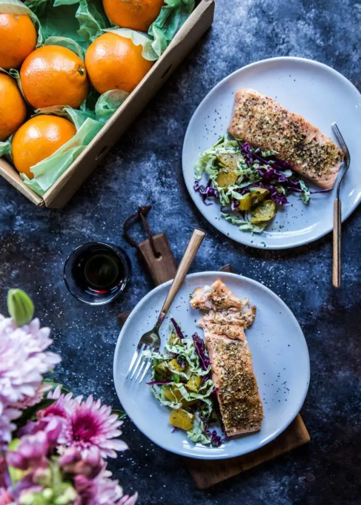 A photo of a salmon recipe with two plates of cooked salmon with slaw next to a box of oranges.