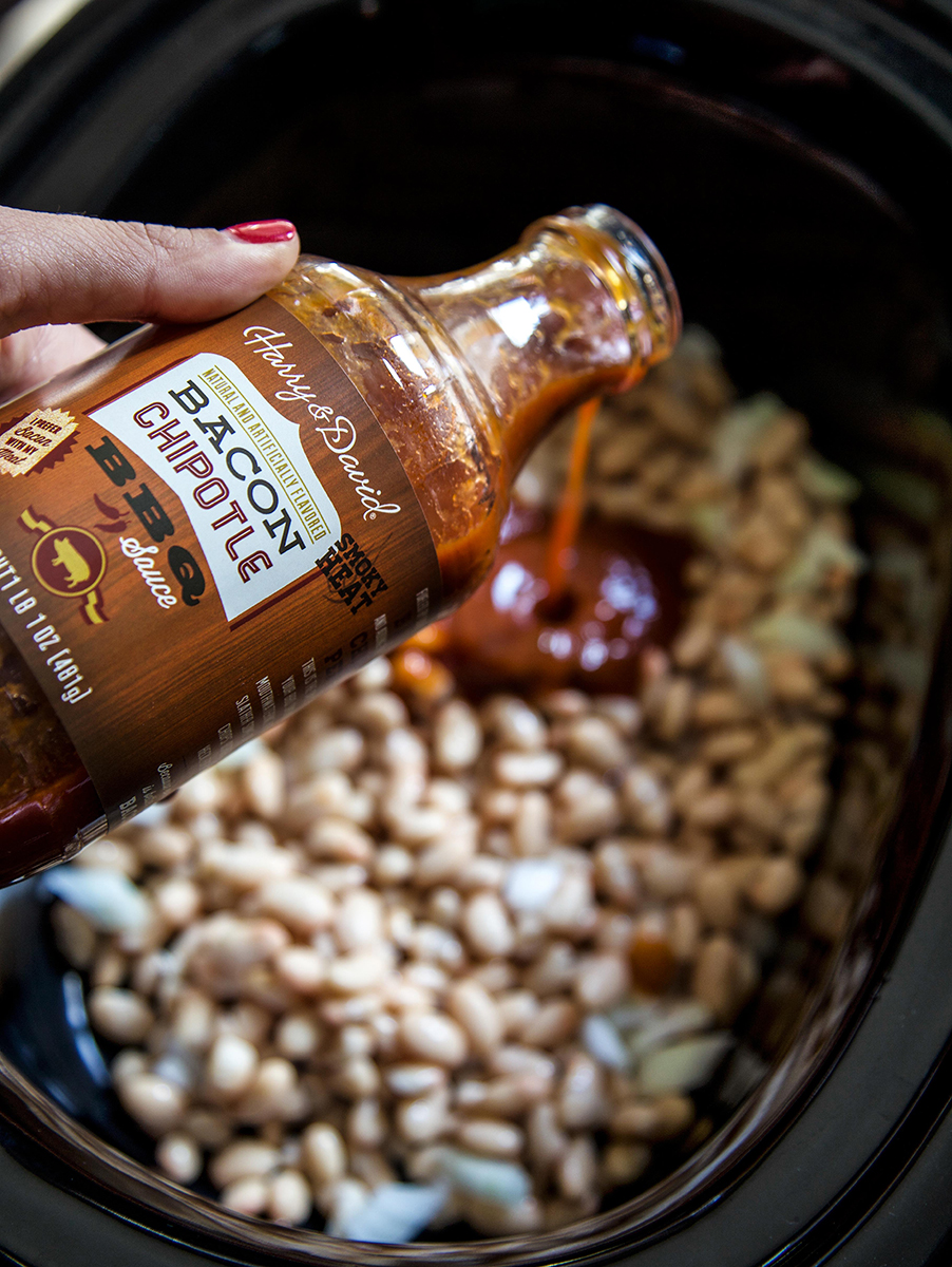 A photo of baked beans recipe with a bottle of barbecue sauce being poured into a pot of beans.