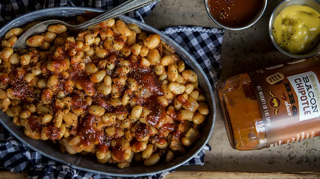 A photo of baked beans recipe with a bowl of baked beans next to a bottle of bbq sauce