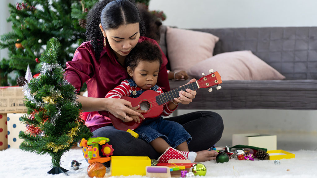 merry christmas in different languages image - mother with son in her lap playing ukulele with christmas decorations in the background. 