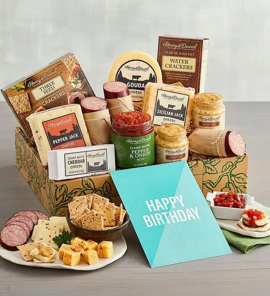 A photo of birthday gift ideas with a box full of cheese, meat and other snacks with a plate of the same items in front of the box next to a card that says, 'happy birthday'.