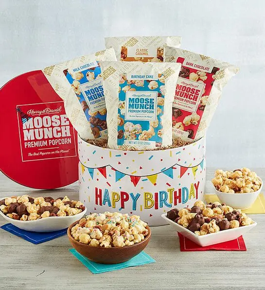 A photo of birthday gift ideas with a tin that has the words "happy birthday" written on the front full of bags of Moose Munch. The same Moose Munch arranged in bowls in front of the tin.