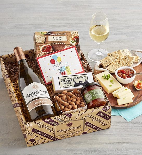 A photo of birthday gift ideas with a box of nuts, cheese and crackers with a bottle of wine and a plate of the same snacks next to the box with a glass of wine.
