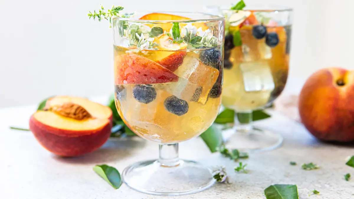 Peach sangria in two glasses surrounded by whole and sliced peaches.