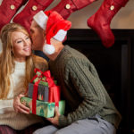 A photo of holiday gifts for him with a woman giving a man a gift while he kisses her cheek