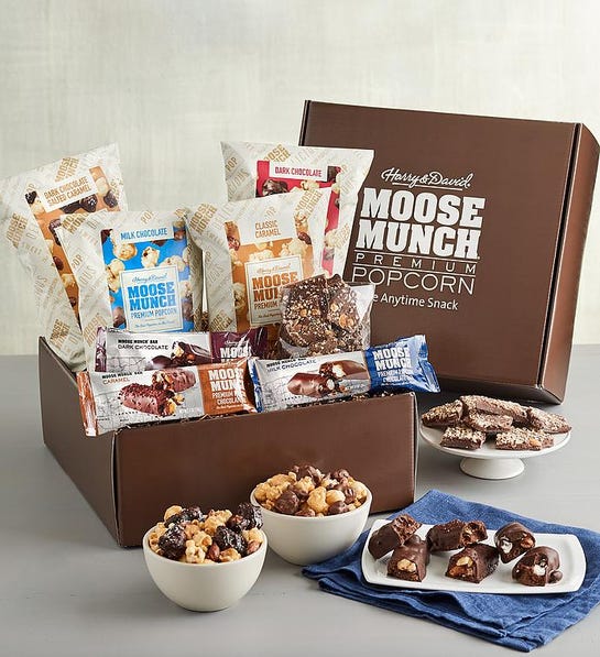 A photo of stocking stuffer ideas with a box of Moose Munch popcorn, bars, and chocolate bark, with bowls and plates in front of the box holding the same items.