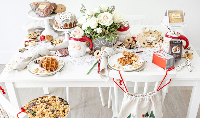 A photo of Christmas crafts for kids with a table full of cookies, cakes, hot chocolate, and waffles with a bouquet of flowers in the middle.