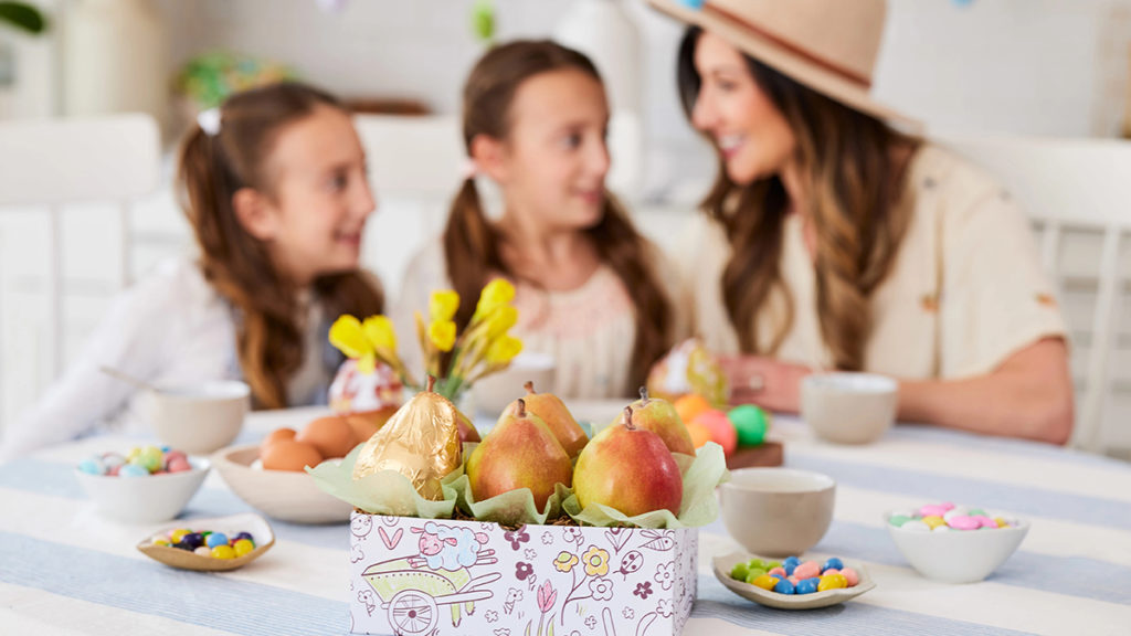 A photo of Easter gift ideas with a box of pears and other Easter candy on a table with a woman and two girls in the background.