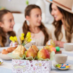 A photo of Easter gift ideas with a box of pears and other Easter candy on a table with a woman and two girls in the background.