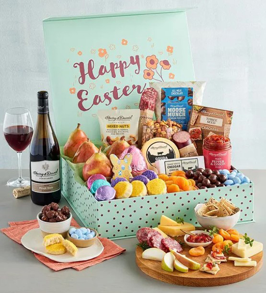 A photo of Easter gift ideas with a box full of fruit, cheese, meat, and other snacks with a bottle of wine next to it and a cutting board full of the same ingredients in front of the box.