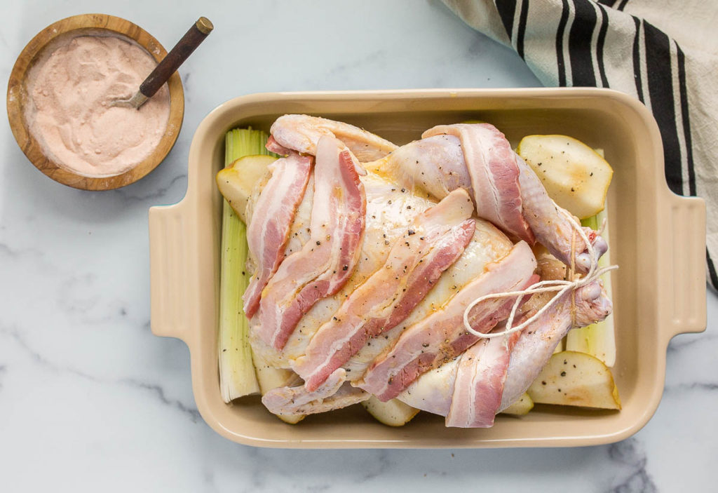 A photo of Thanksgiving chicken with a raw chicken in a baking pan covered in bacon with a bowl of salt next to it