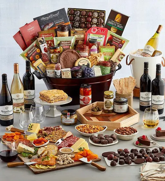 A photo of a gift guide with a basket overflowing with charcuterie, cheese, cake, and other snacks with a spread of the same items in front of the basket with six bottles of wine surrounding it.