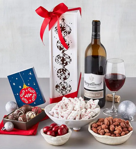 A photo of a gift guide with a bottle of wine and a full glass sitting behind several bowls of nuts and chocolate covered fruit and nuts with an open box of truffles next to them.