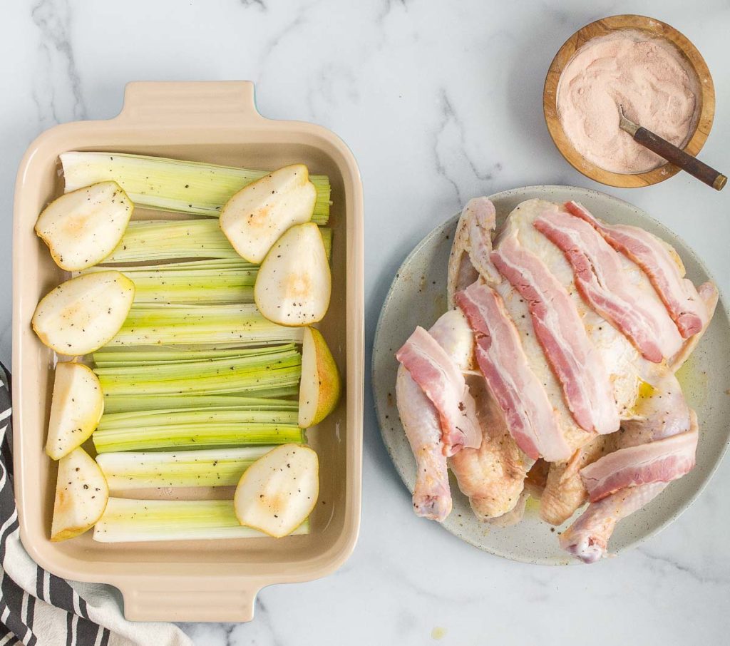 A photo of Thanksgiving chicken with a raw chicken covered in a bacon on a plate next to a baking pan full of pears and celery