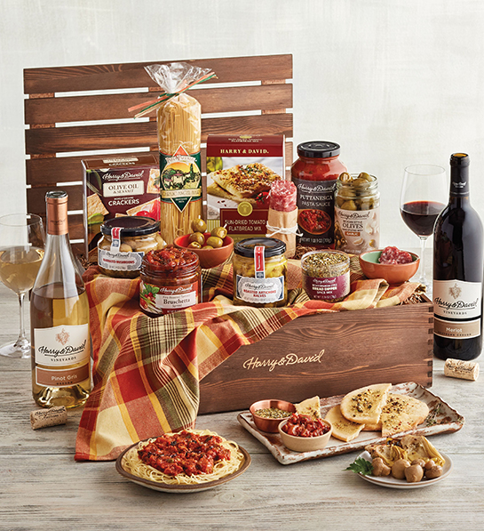A photo of gift baskets with a wooden box filled with dried pasta, jars of tomato sauce and other antipasto bites with two bottles of wine next to it and plates full of food in front