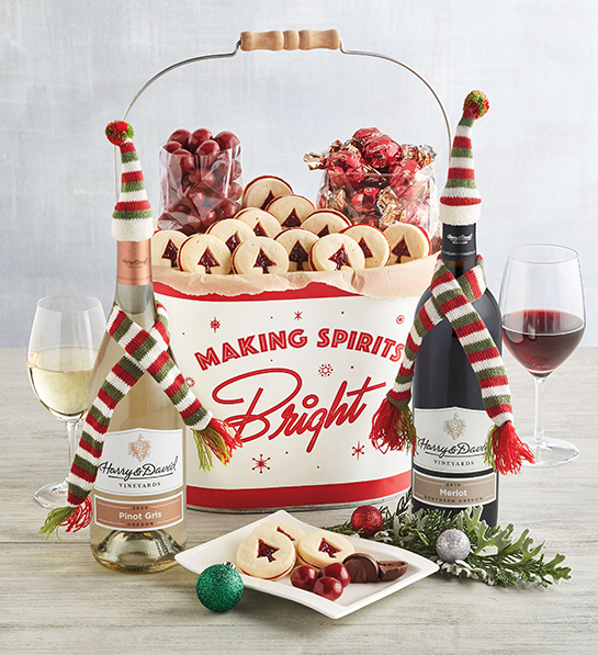 A photo of gift baskets with two bottles of wine wrapped in tiny scarves and hats with a tin bucket of cookies and a glass of wine behind them