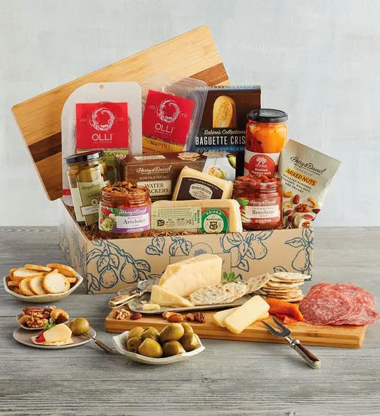 A photo of gift baskets with a box full of cheese, jars of olives and spreads, and crackers with a cutting board in front full of cheese and crackers.