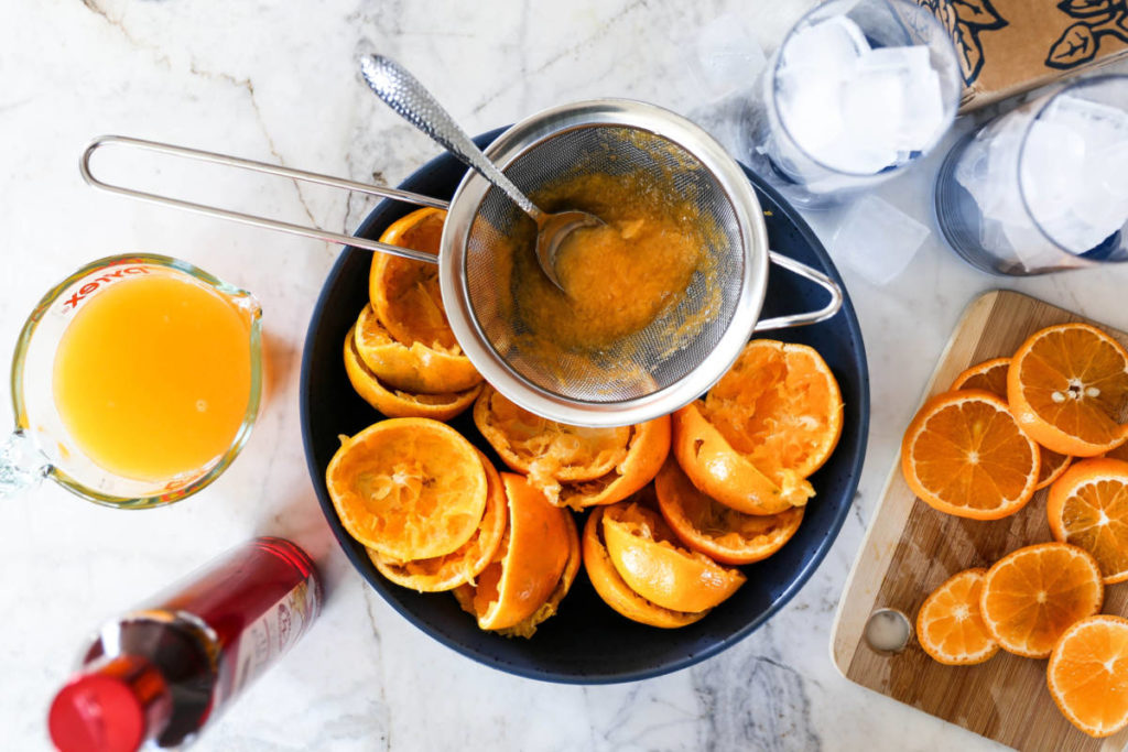 A photo of a mocktail recipe with lots of oranges cut in half and being squeezed through a strainer