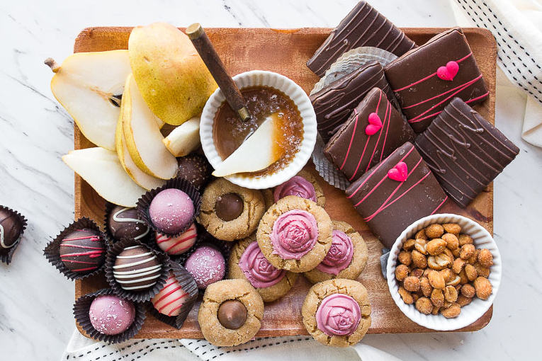 A photo of Valentine's Day date ideas with a board full of cookies, pear slices, and truffles with a ramekin of caramel dipping sauce in the middle.