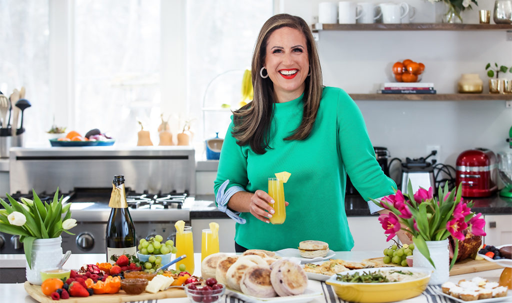 A photo of Easter meal with a woman holding a mimosa standing in front of an Easter brunch spread