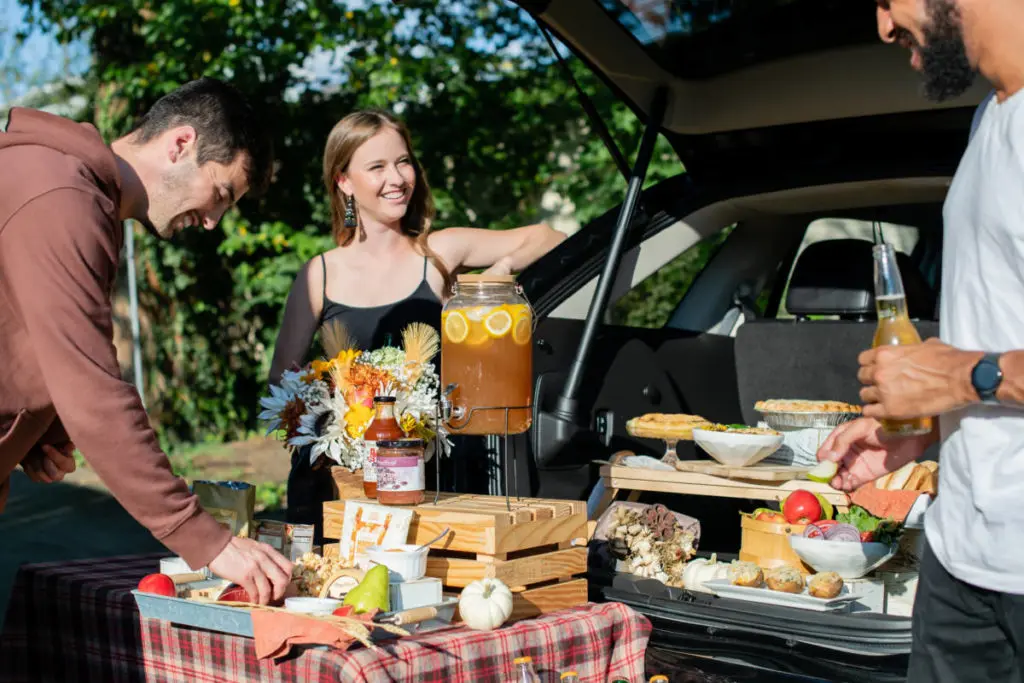 A photo about a tailgate party. A woman leaning against a car talking to two men with a display of snacks and drinks in front her.