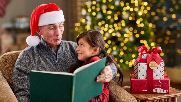 A photo of Christmas gifts with a granddaughter sitting in her grandfather's lap while he reads to her with Christmas decorations in the background.