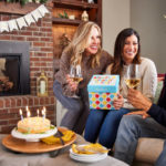 A photo of december birthdays with three people sitting on a couch drinking wine and opening a birthday present