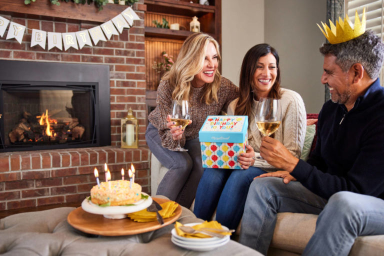 A photo of december birthdays with three people sitting on a couch drinking wine and opening a birthday present