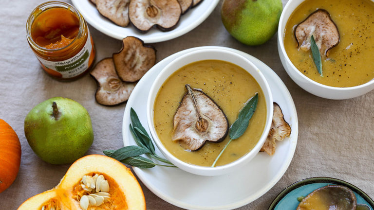 A photo of pumpkin soup in a bowl with a fresh pear, a bowl of dried pears, and a pumpkin surrounding it