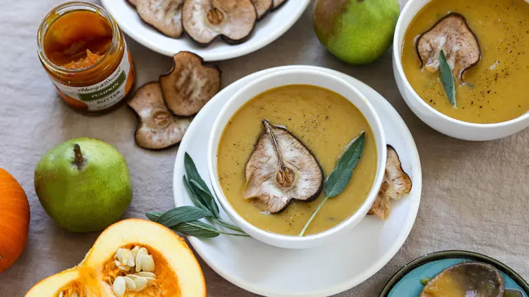 A photo of pumpkin soup in a bowl with a fresh pear, a bowl of dried pears, and a pumpkin surrounding it
