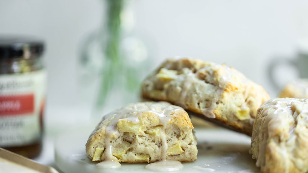 A photo of an apple scone recipe with three scones stacked on each other and a jar of apple butter in the background