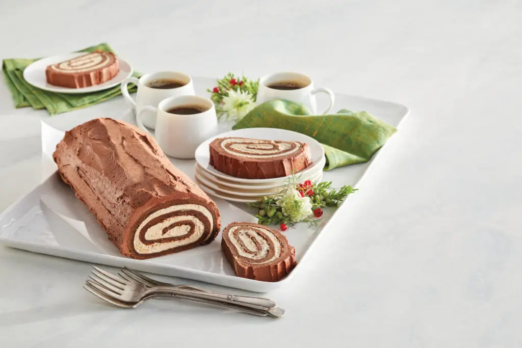 A photo of a bûche de Noël on a platter surrounded by cups of coffee and several slices of the bûche de Noël on plates.