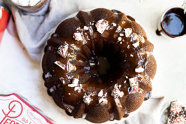 A photo of a bundt cake decorated with frosting and bits of peppermint bark sitting next to a tin of peppermint bark and a cup of coffee