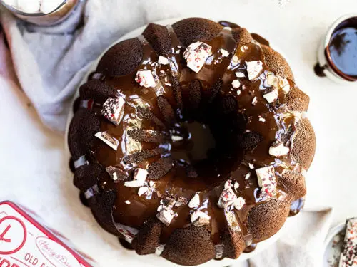 A photo of a bundt cake decorated with frosting and bits of peppermint bark sitting next to a tin of peppermint bark and a cup of coffee