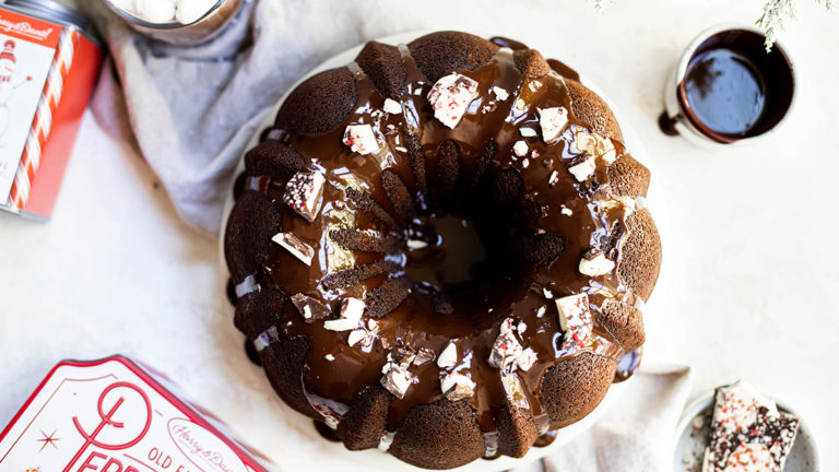 A photo of a bundt cake decorated with frosting and bits of peppermint bark sitting next to a tin of peppermint bark and a cup of coffee