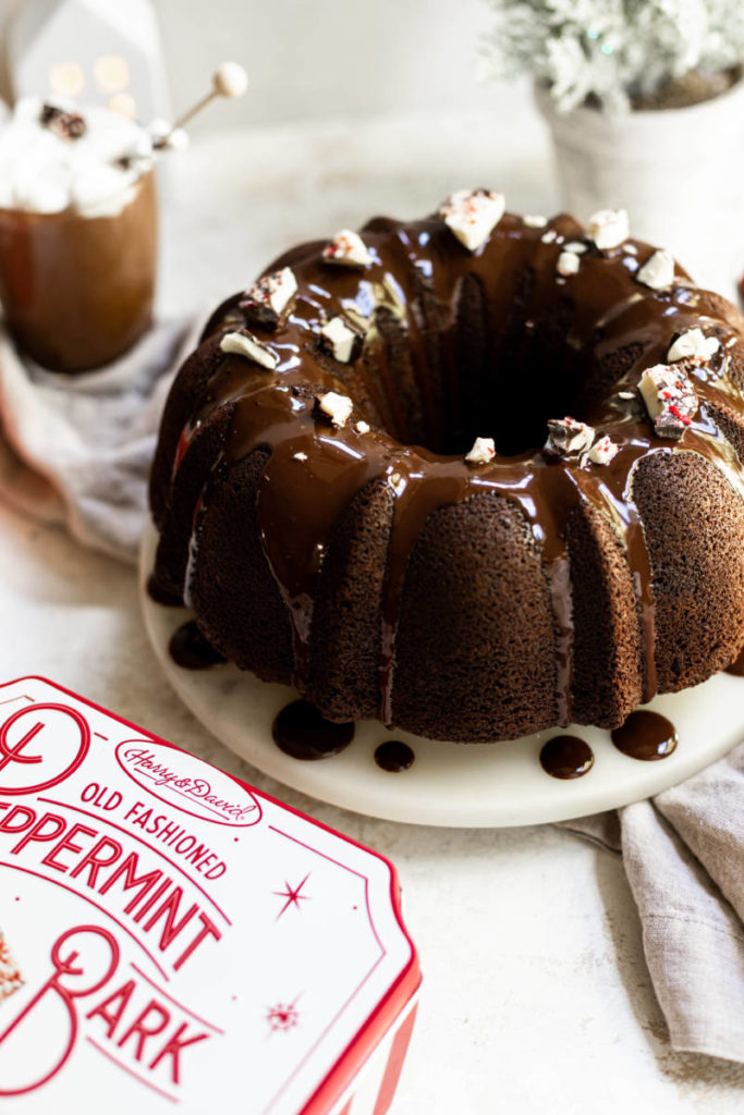 A photo of a frosted bundt cake sitting next to a tin of peppermint bark and a cup of hot chocolate
