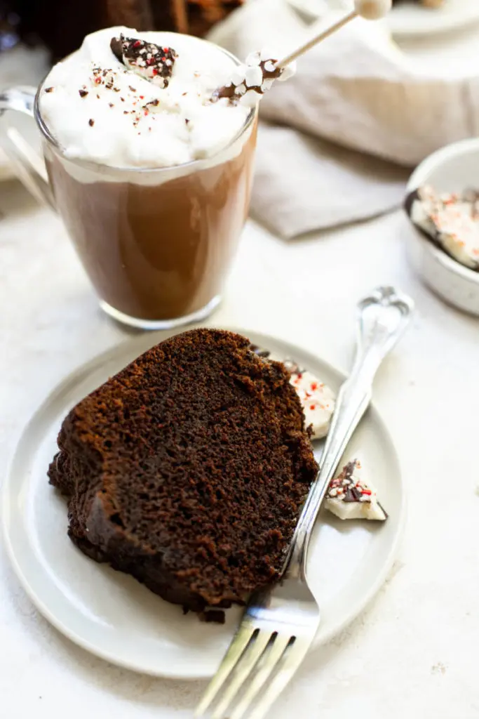 A photo of a bundt cake with a slice of it on a plate next to a cup of hot chocolate