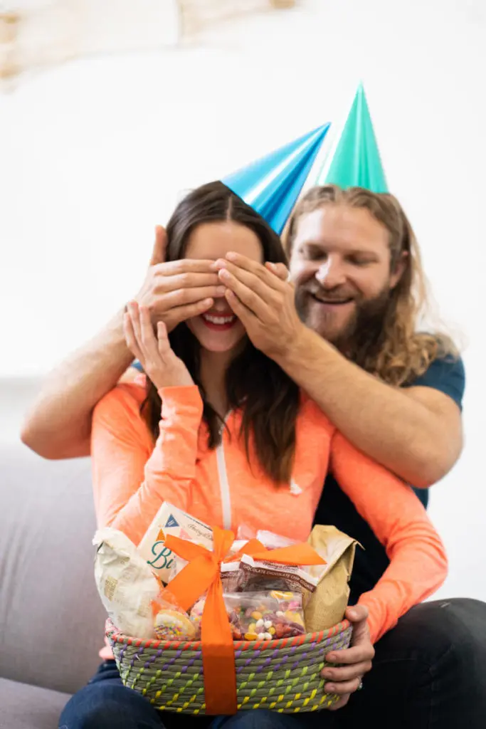 A photo of december birthdays with a woman sitting next to a man who is covering her eyes so she can't see the present in her lap