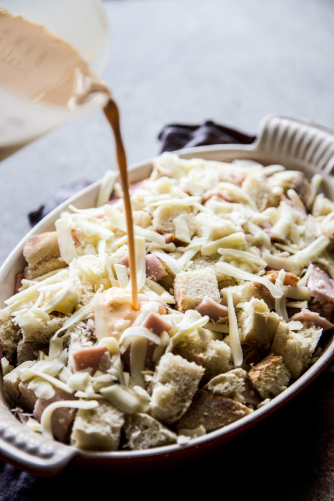 A photo of an eggs benedict casserole with someone pouring liquid into a casserole dish that's full of bread and cheese